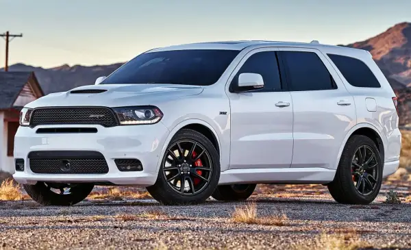 Biały Dodge Durango 3.6 SRT z czarnymi felgami i czerwonymi zaciskami hamulcowymi stoi na żwirowej drodze, w tle góry i opuszczony dom.