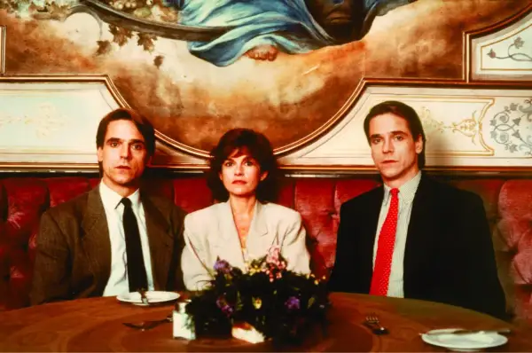 Jeremy Irons w filmach, tutaj z dwiema innymi osobami, siedzi przy stole w eleganckiej restauracji.