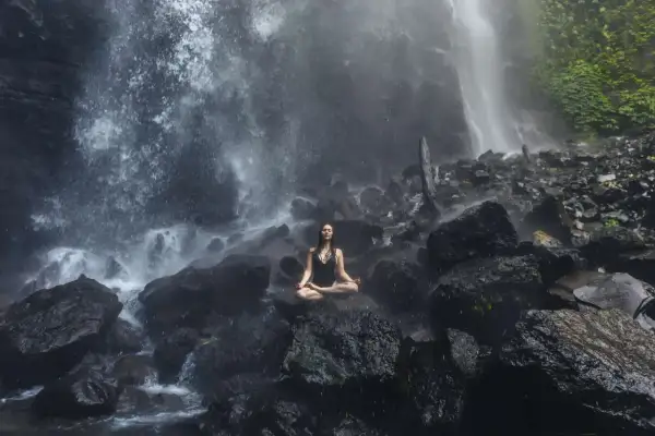 Mujer meditando en posición de loto sobre rocas, rodeada por la bruma de una cascada. Un momento de sanación pránica en la naturaleza.