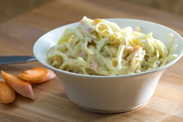 En skål med krämig, godaste coleslaw med strimlad vitkål och morötter. Bredvid ligger några morotsskivor.