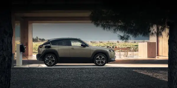 Mazda MX-30 ładuje się przy nowoczesnym domu z drewnianymi elementami, w otoczeniu natury.