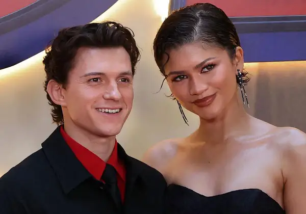 Tom Holland y Zendaya sonríen juntos en un evento. Él lleva una camisa roja y corbata negra. Ella luce un vestido negro y pendientes largos.