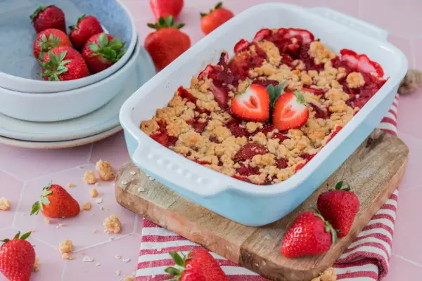 Ein frisch gebackener Erdbeer Crumble in einer hellblauen Auflaufform, garniert mit Erdbeerhälften. Daneben eine Schüssel mit Erdbeeren und verstreute Streusel.