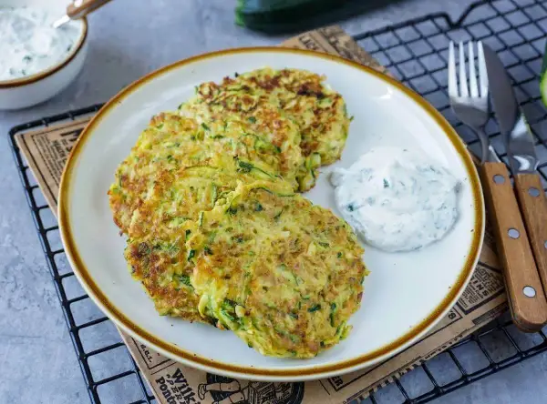 Drei goldbraune Zucchini-Pfannkuchen liegen auf einem Teller, daneben ein Klecks Kräuterquark. Ein leckeres Gericht!
