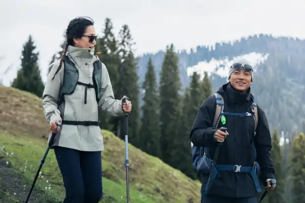 Para w odpowiednim ubraniu w góry, z kijkami trekkingowymi, cieszy się wędrówką pośród zielonych wzgórz i ośnieżonych szczytów.
