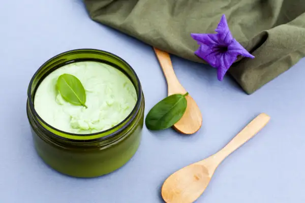 Un pot de gommage cuir chevelu maison, vert pâle, avec des cuillères en bois et une fleur violette.