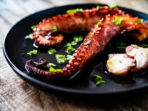 Tentacules de poulpe grillés, parsemés de persil frais, sur une assiette noire. La cuisson du poulpe révèle sa texture tendre.