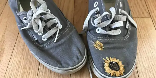 Paire de chaussures grises avec des lacets blancs. L'une des chaussures est ornée de broderies de tournesols, donnant un air bohème à ces chaussures trouées.