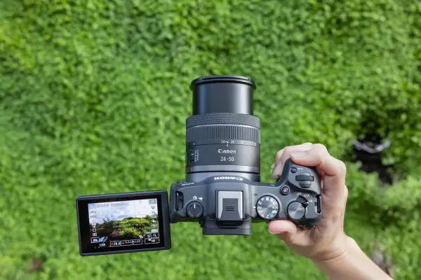 En hand håller en Canon systemkamera med objektivet 24-50mm. Kamerans skärm visar en grönskande vy.