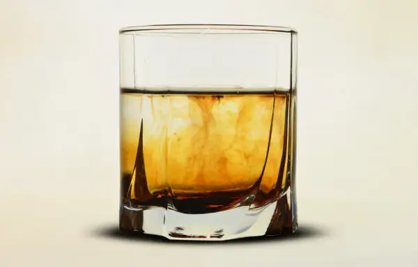 Szklanka z bursztynowym płynem, w którym rozpuszcza się ciemniejsza substancja, przypominająca proces, jak zrobic whiskey.