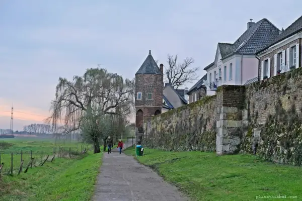 Ein Paar spaziert auf einem Weg entlang der alten Stadtmauer von Zons am Rhein. Ein Weidenbaum und ein Wehrturm prägen die Szenerie.