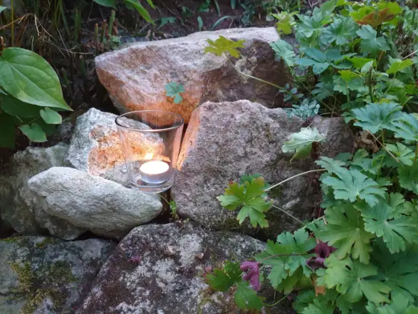Ein Teelicht in einem Glas leuchtet zwischen Steinen und grünen Pflanzen. Ein Symbol für Hoffnung und einen bibelvers mut.