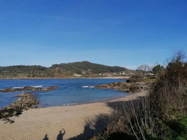 La tranquila playa de Vilariño, con arena dorada y aguas azules, se extiende bajo un cielo despejado. Rocas salpican la orilla, y colinas verdes cubiertas de árboles la rodean.