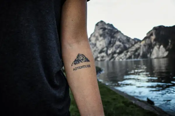 Tatuaż góry z napisem "ADVENTURE" na ramieniu, na tle jeziora i skalistych szczytów.