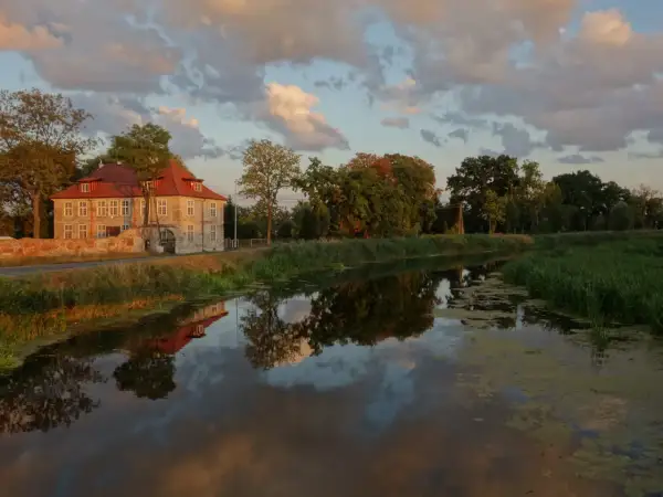 Dworek w Gdańsku Olszynce z czerwonym dachem odbija się w spokojnej wodzie kanału. Chmury na niebie malują zachód słońca.