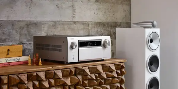 Srebrny amplituner Denon z wyświetlaczem Dolby Atmos, obok białej kolumny głośnikowej. To centrum domowego kina, które pozwala cieszyć się kinową jakością dźwięku.