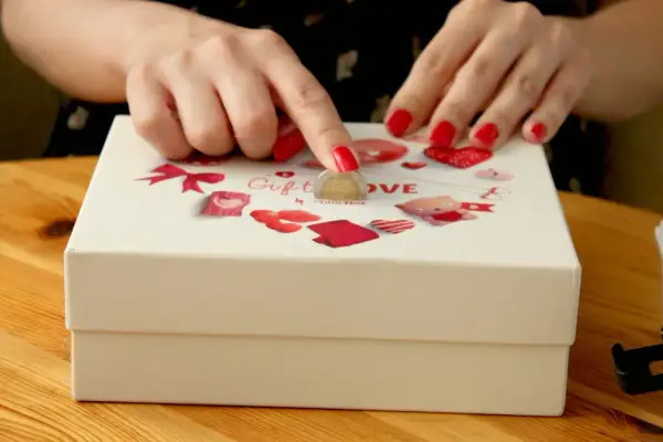 Dłonie z czerwonymi paznokciami wkładają monetę do pudełka z napisem "Gift LOVE".