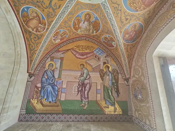 Mozaika przedstawia scenę biblijną z postaciami w stylu sztuki sakralnej. Widać Matkę Boską, anioła i mężczyznę, a także symbole religijne.