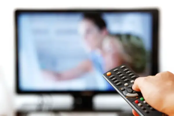 Dłoń trzymająca pilot do telewizora przed ekranem.