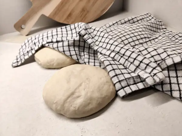 Två runda pizzadegar ligger på en bänk, en täckt av en rutig kökshandduk. Redo för bakning samma dag.