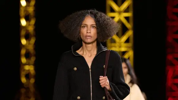 Una mujer con cabello rizado y gris camina por una pasarela, luciendo un elegante cárdigan negro con botones dorados, como una **chanel cantante**.