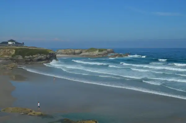 La **playa Os Castros** se extiende bajo un cielo azul, con olas rompiendo en la arena dorada. Una casa moderna corona el acantilado.
