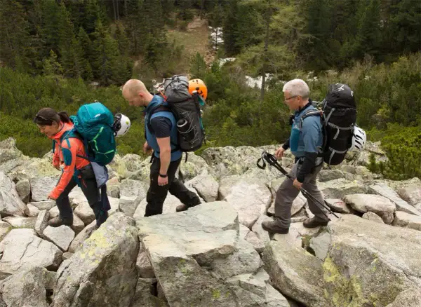 Drei Wanderer mit Rucksäcken und Helmen auf felsigem Untergrund. Die **Packliste Klettersteig** ist gut gefüllt für das Abenteuer.