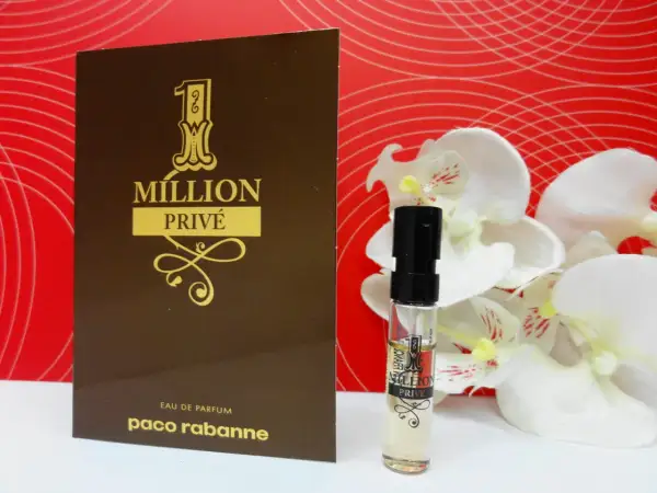 Złoty flakonik i próbka Paco Rabanne 1 Million Privé na tle czerwonych fal i białych kwiatów.