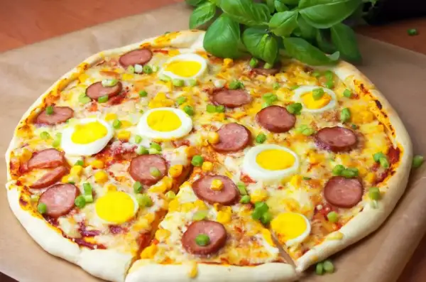 Pyszna pizza z jajkiem, kiełbasą i kukurydzą, ozdobiona zieloną cebulką i bazylią.