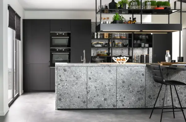 Cuisine moderne aux façades grises et à l'îlot central en sol terrazzo. Électroménagers intégrés et étagères noires remplies d'objets décoratifs.