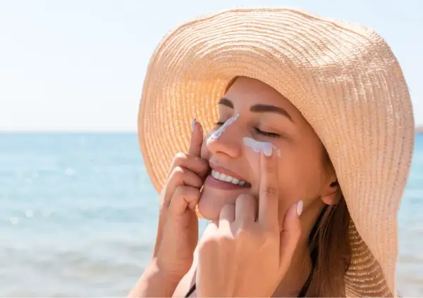 Une femme souriante applique de la crème solaire sur son visage, protégeant sa peau. Est-ce que la crème solaire hydrate ? Oui, certaines le font !