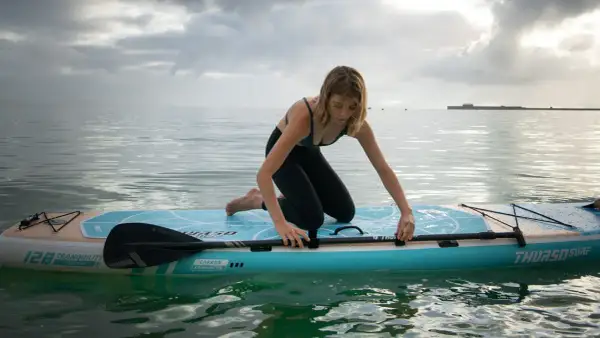 Donna in ginocchio su un sup, pronta a pagaiare. La tavola galleggia su acqua calma sotto un cielo nuvoloso.