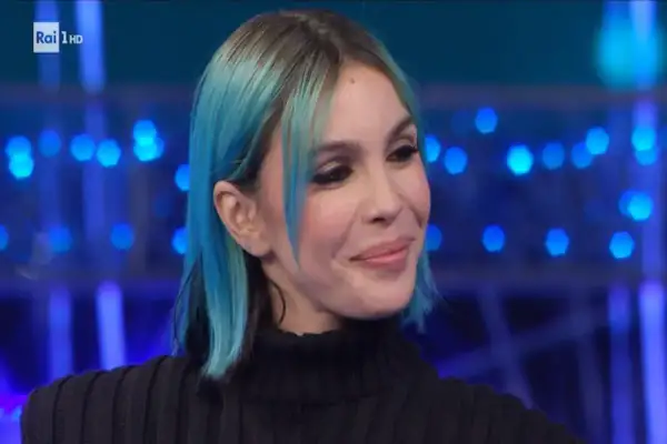 Una donna con capelli blu e neri, trucco marcato, sorride con un'espressione da rose villain.