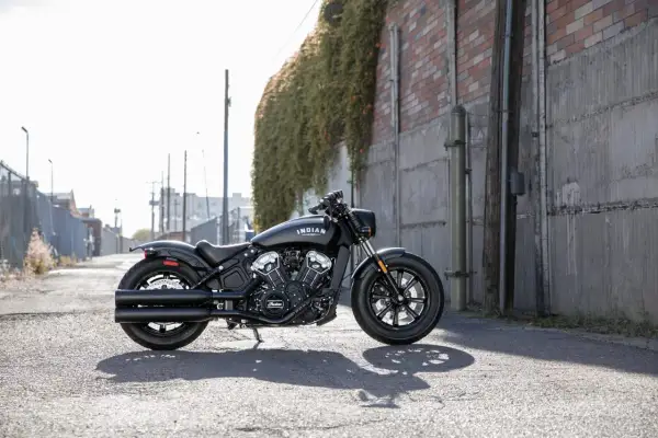 Czarny motocykl cruiser Indian Scout stoi na ulicy, otoczony miejską scenerią z ceglanym murem i zielenią.