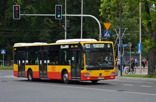 Żółto-czerwony autobus linii 103 w Warszawie jedzie ulicą. W tle drzewa i sygnalizacja świetlna.