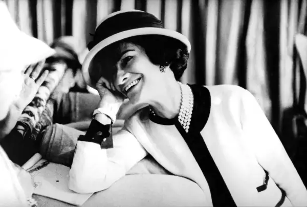 Uśmiechnięta kobieta w kapeluszu i perłach, z dłonią opartą o policzek. Ikona stylu, Coco Chanel, w swoim charakterystycznym, eleganckim stroju.