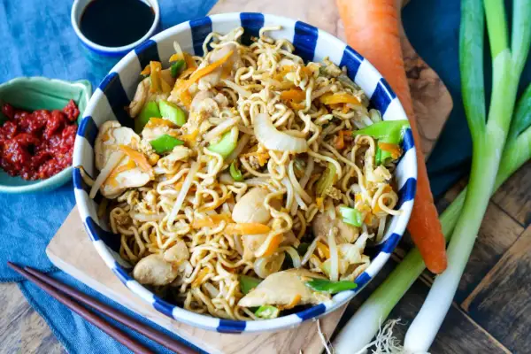 Bami Goreng mit Hühnchen, Karotten und Frühlingszwiebeln in einer Schüssel, daneben Sojasauce, Chili und frisches Gemüse.