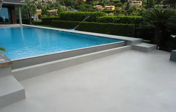 Modern villa with a bright blue pool and surrounding terraces finished with elegant piscinas de microcemento. Lush green hedges and palm trees frame the scene.
