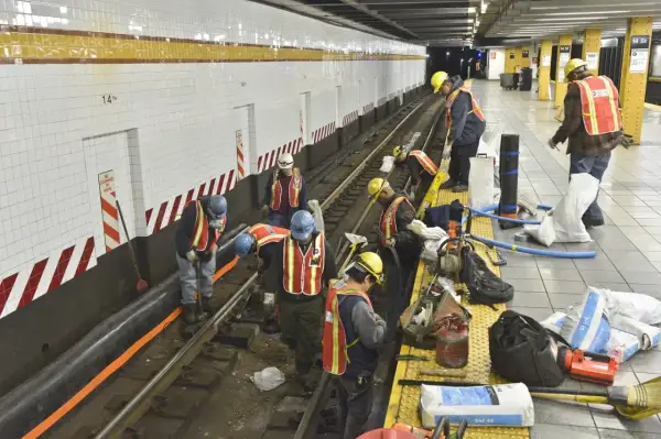 Des ouvriers en tenue de chantier, gilets de sécurité et casques, travaillent sur les voies d'une station de métro. Ils semblent engagés dans des travaux de maintenance, peut-être le prime habillage ou deshabillage de certains éléments.