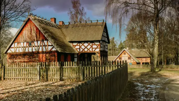 Drewniane domy z krytymi strzechą dachami i drewnianym płotem na tle drzew.