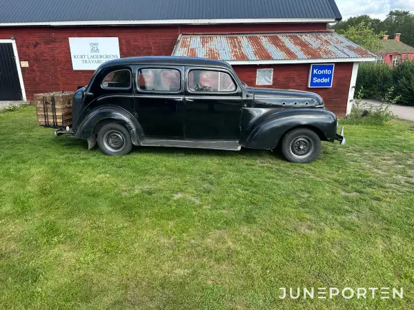 Svart Volvo PV 831 parkerad på gräs framför en röd byggnad med rostigt plåttak. En träkorg sitter på bakre stötfångaren.