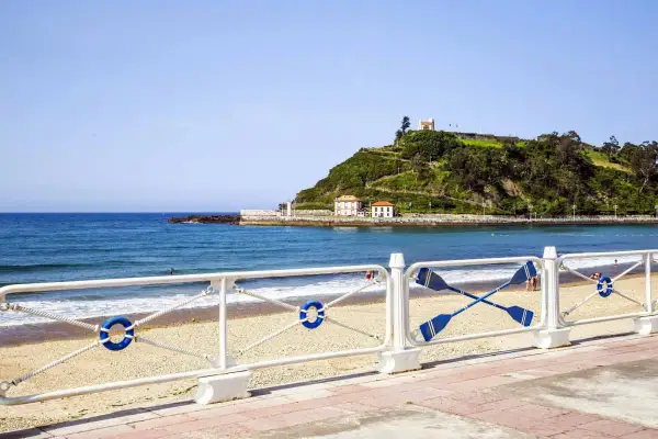 La **playa de Santa Marina** con su arena dorada, el mar azul y una colina verde coronada por un edificio blanco.