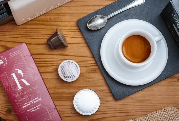 Una tazzina di espresso fumante, capsule e confezione Rossini: un invito a gustare una nota varietà di caffè.