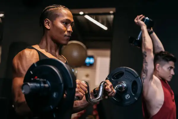 Zwei Männer trainieren im Fitnessstudio. Der vordere Mann hebt eine Hantel, der hintere Mann stemmt eine Kurzhantel. Verschiedene Kraft Arten werden hier gezeigt.