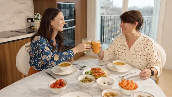 Deux femmes trinquant avec des jus verts et orangés, entourées d'un repas sain. Un exemple de comment ne pas grossir à la ménopause.