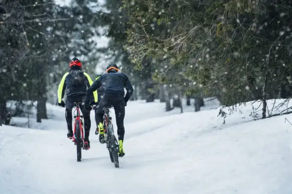 Dwóch rowerzystów przemierza zimowe trasy rowerowe Olsztyn, podczas gdy śnieg delikatnie opada na las.