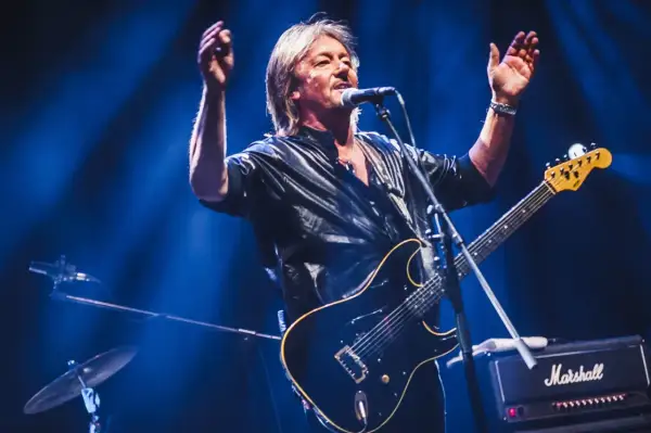 Chris Norman, lider zespołu Smokie, śpiewa i gra na gitarze na scenie, oświetlony niebieskim światłem.