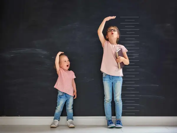 Due bambine misurano la loro altezza su una lavagna. Una, con un'espressione sorpresa, sembra avere l'altezza di un bambino di 2 anni.
