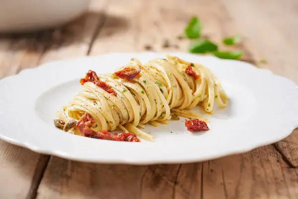 Un piatto di linguine condite con pomodori secchi, capperi e prezzemolo, su un piatto bianco con sfondo in legno.