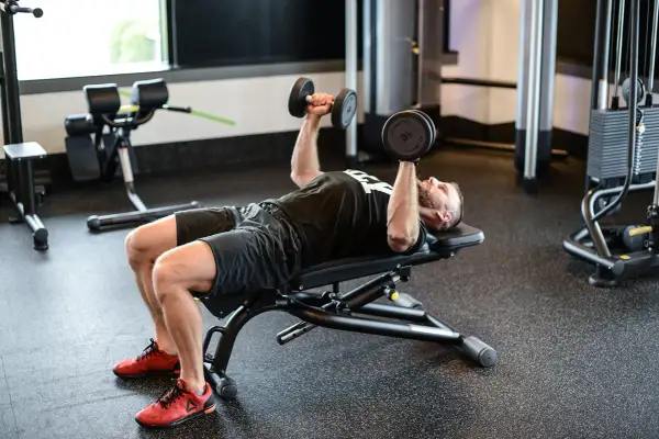 Mężczyzna wykonuje ćwiczenie dumbbell press na ławce w siłowni, podnosząc ciężarki nad klatką piersiową.